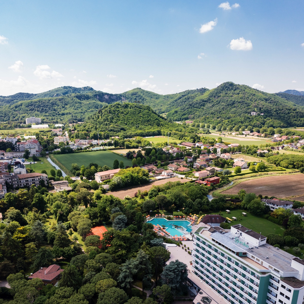Veduta aerea di resort termale con hotel e piscina tra colline verdi, campi e borgo rurale sotto cielo azzurro.