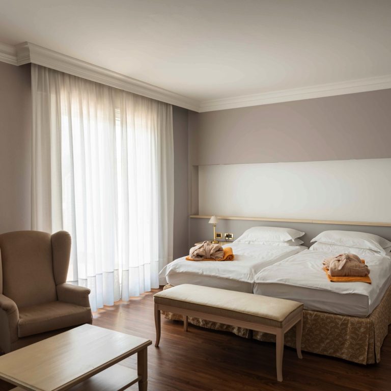 Camera d’hotel luminosa con due letti singoli, poltrona e panca, parquet e tende bianche, stile elegante e accogliente.