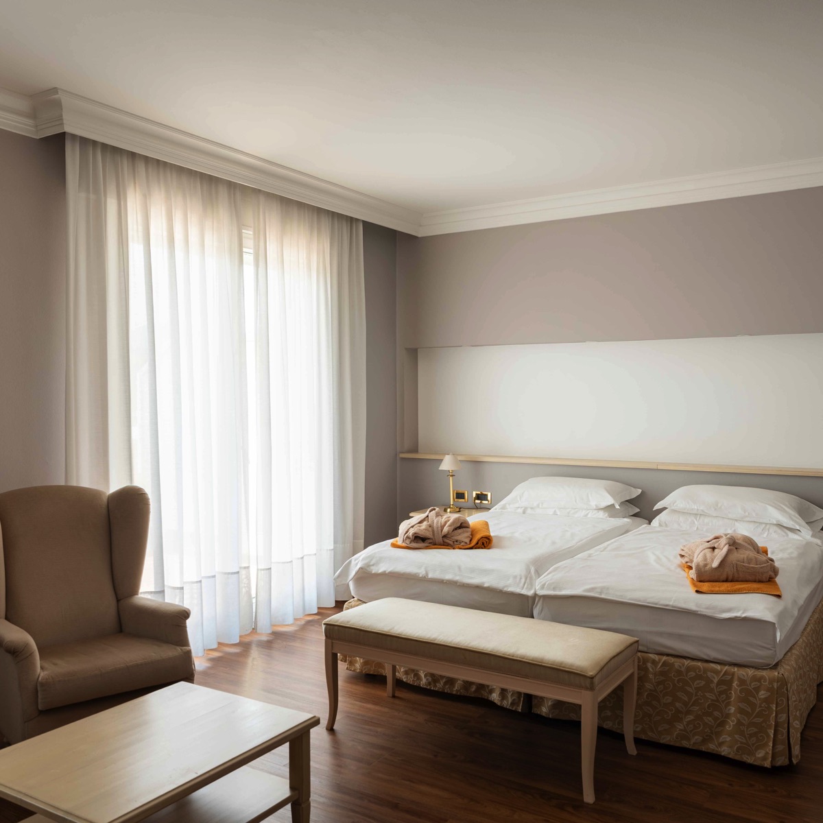 Camera d’hotel luminosa con due letti singoli, poltrona e panca, parquet e tende bianche, stile elegante e accogliente.