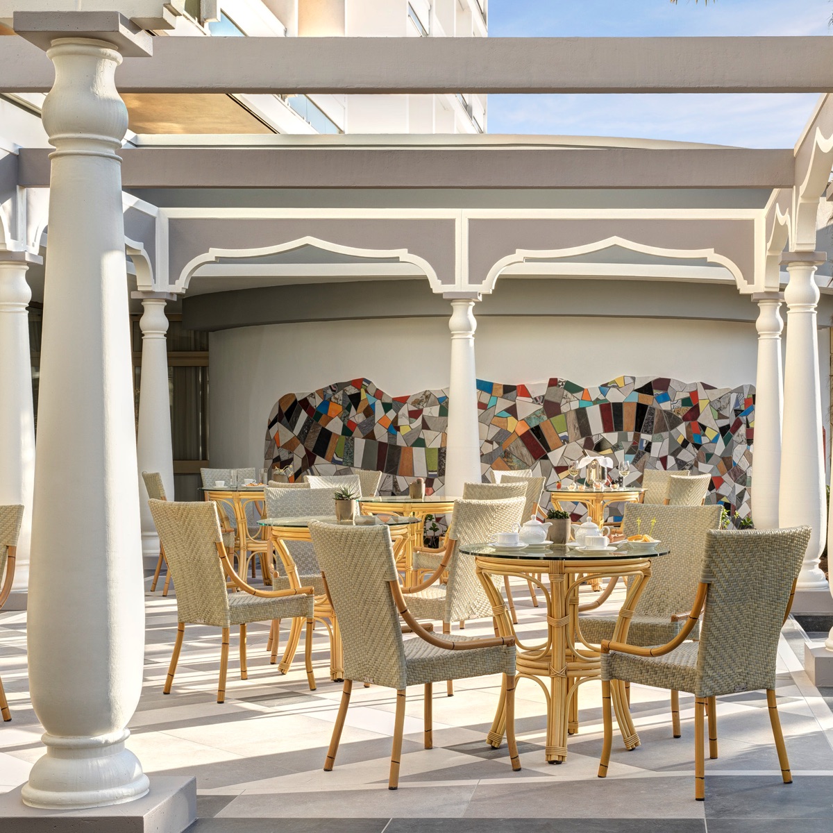 Terrazza ristorante hotel di lusso con pergola, sedie in rattan, tavoli in vetro e parete a mosaico colorato all'aperto