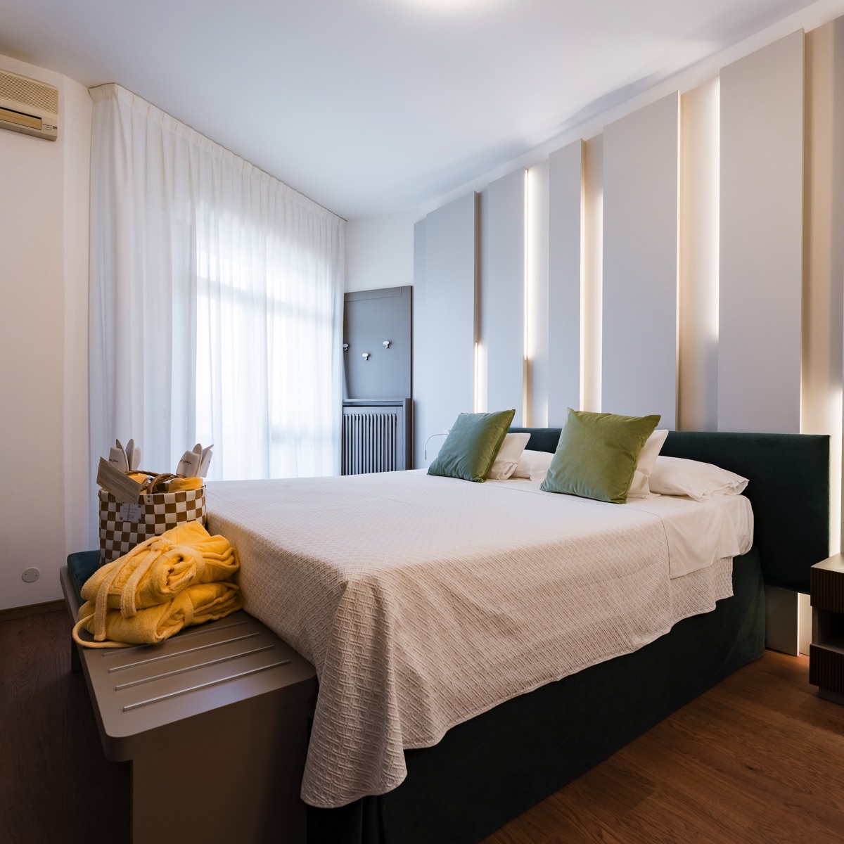 Camera da letto moderna con letto king, testiera a pannelli illuminati, cuscini verdi, tenda bianca e parquet caldo.