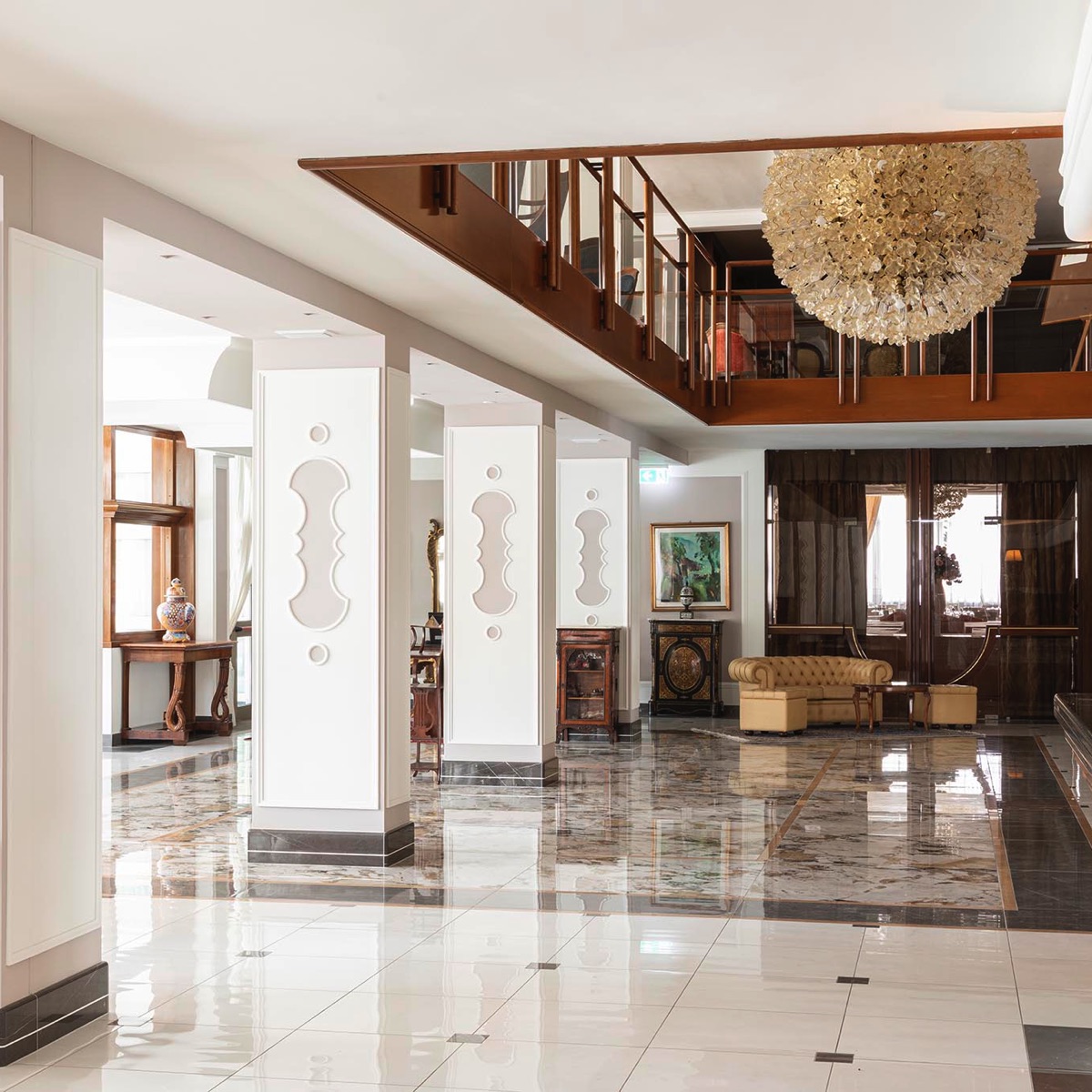 Lobby hotel di lusso con pavimento in marmo, lampadario scenografico, mezzanino in legno e divano beige.