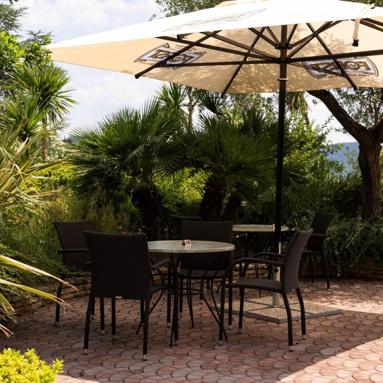 Terrazza giardino con tavolo rotondo e sedie in rattan sotto grande ombrellone, palme e pavimento in pietra, soleggiato