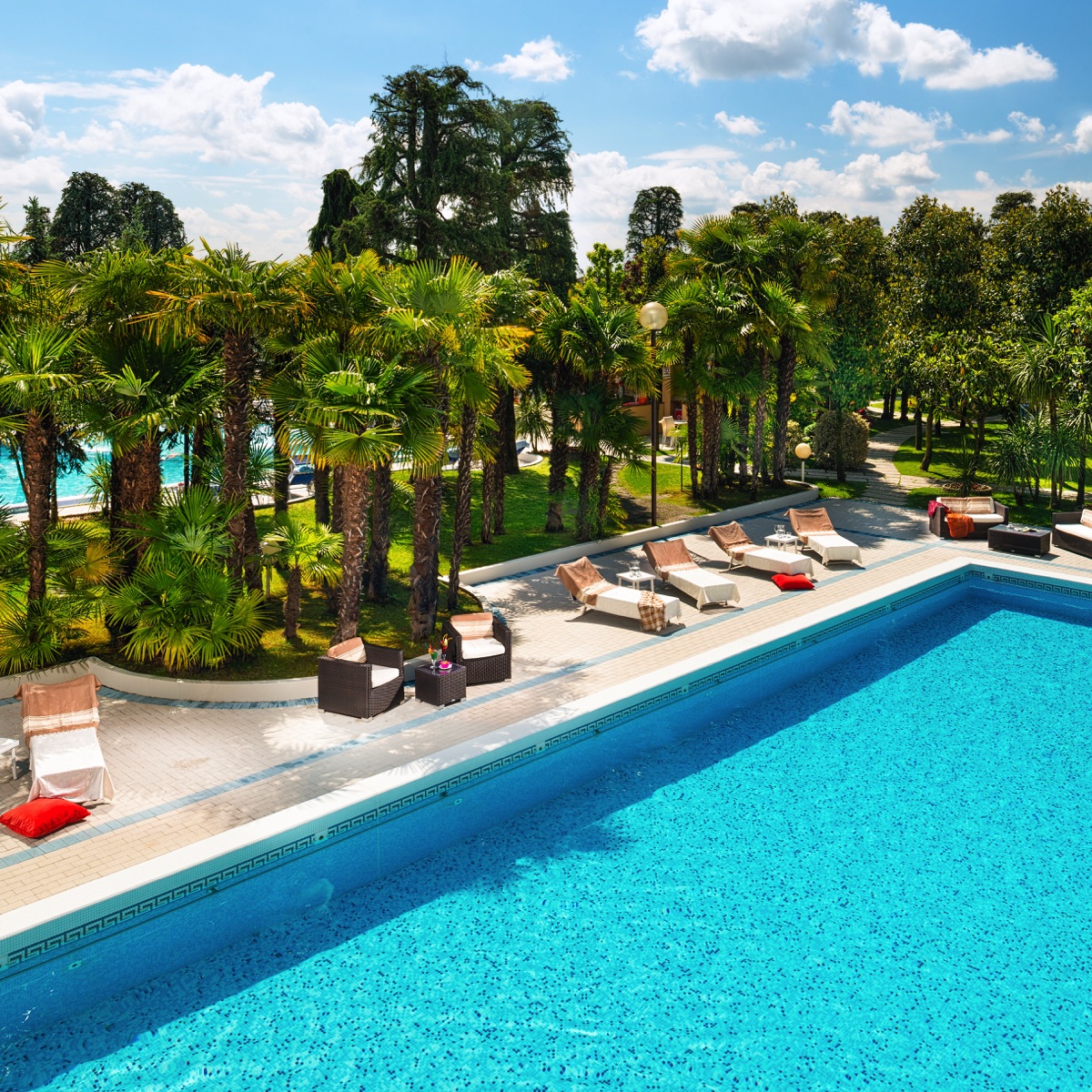 Piscina turchese di resort con lettini e palme, giardino curato e cielo azzurro: relax estivo all'aperto.