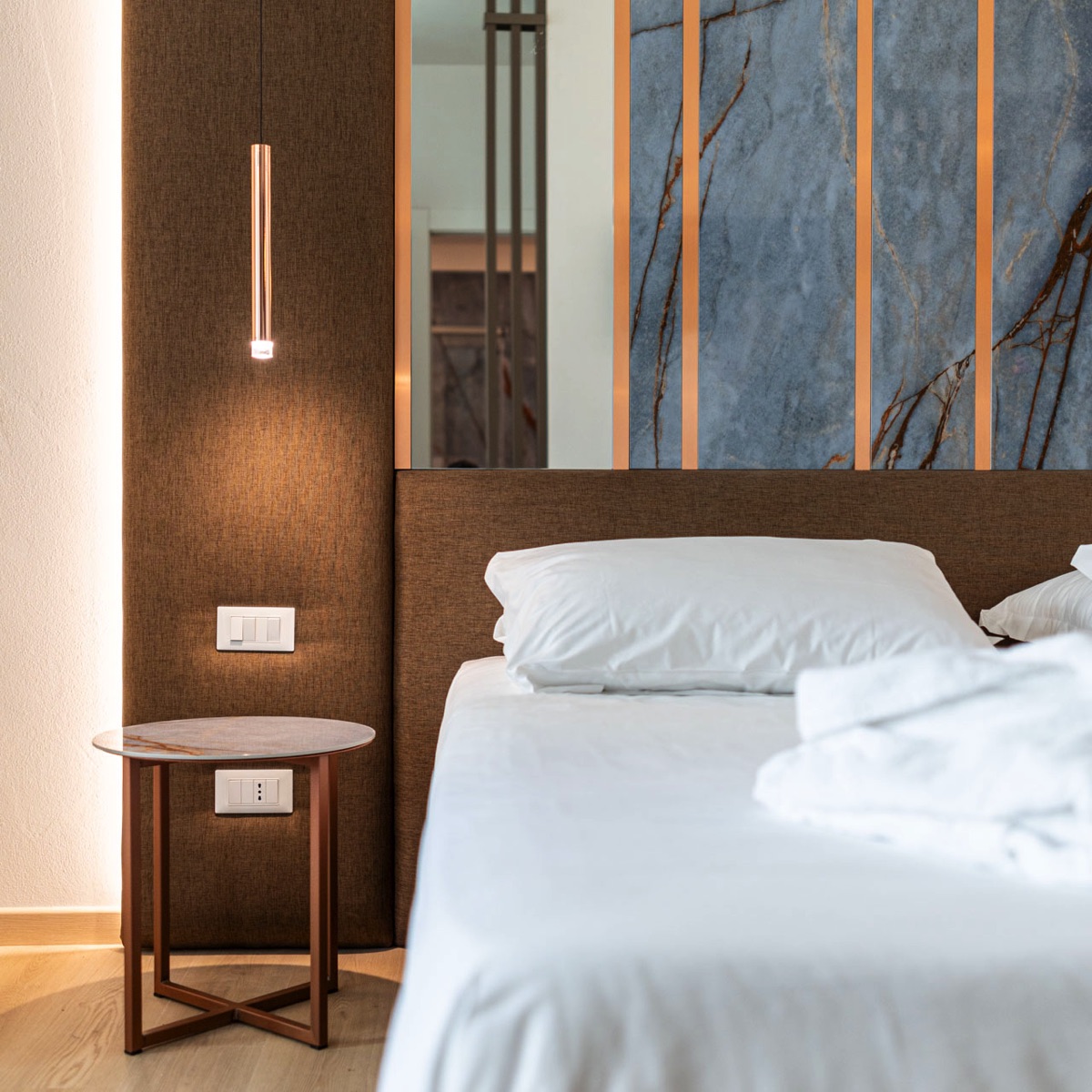 Camera d’hotel design con letto matrimoniale bianco, testata imbottita, lampada a sospensione e pannelli in marmo azzurro