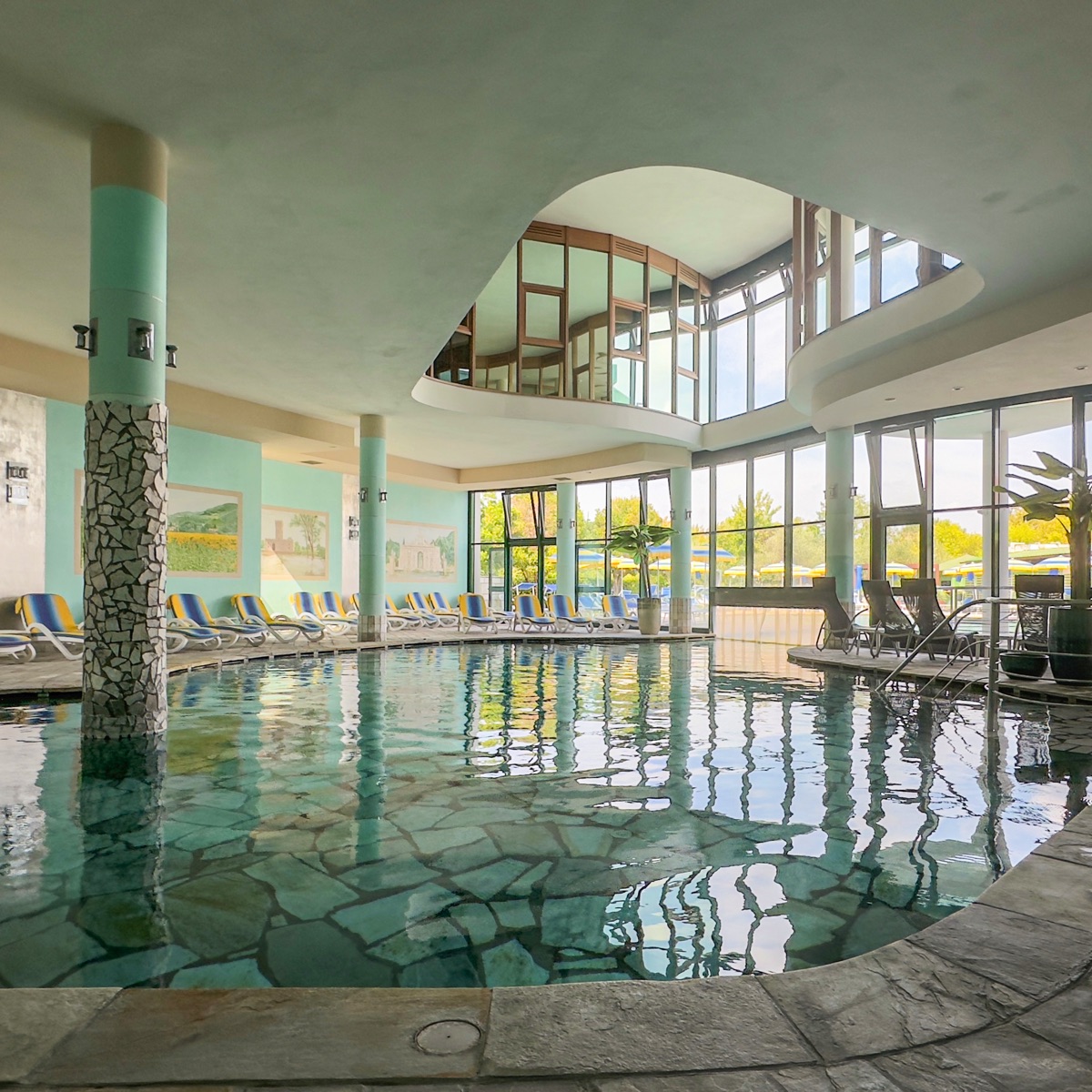 Piscina termale coperta con pavimento in pietra, vetrate luminose e lettini relax in spa moderna con vista sul giardino.