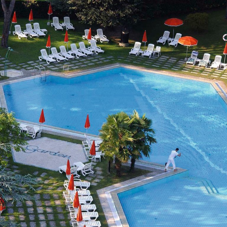 Grande piscina all'aperto dell’hotel con lettini bianchi, ombrelloni arancioni e palme su prato curato, ideale relax