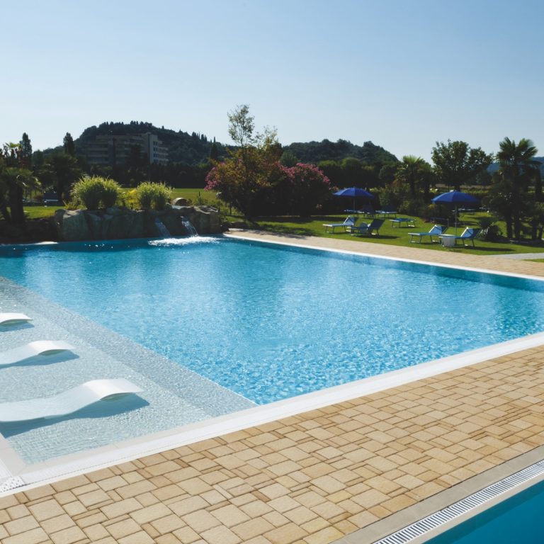 Piscina all'aperto di hotel con lettini in acqua, cascata rocciosa e giardino con ombrelloni, vista sulle colline