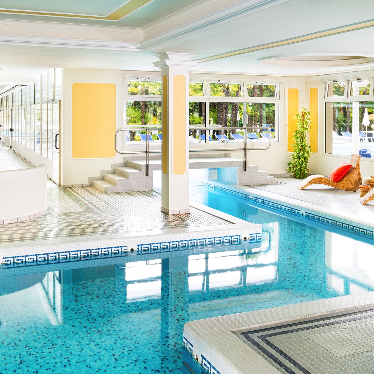 Piscina interna spa luminosa con piastrelle a mosaico, idromassaggio e vista su palme; area relax di hotel benessere.