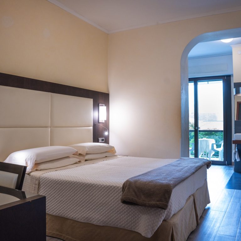 Camera hotel moderna con letto matrimoniale, arredo minimal e balcone luminoso con tavolino, parquet e vista giardino