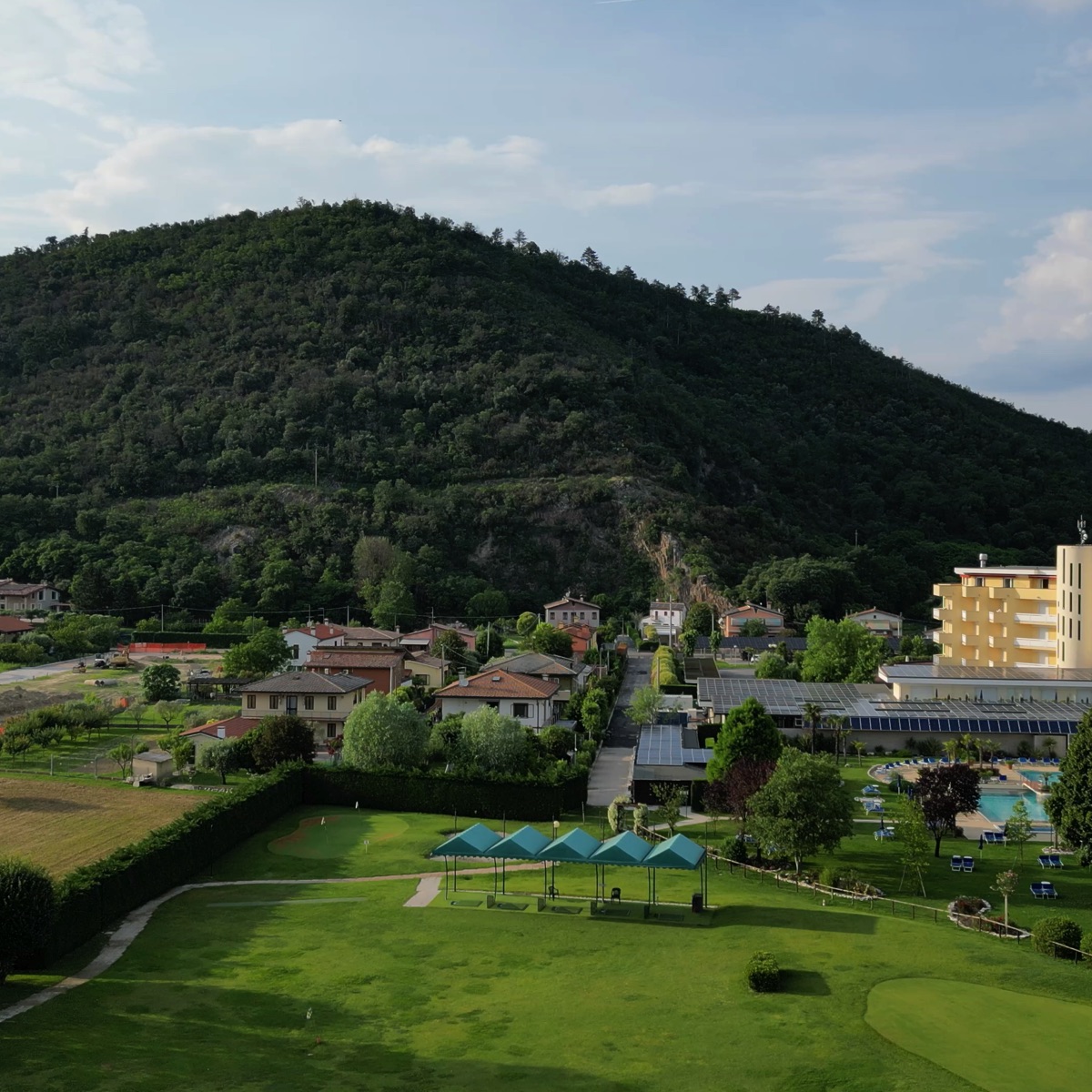 Resort con piscina e campo pratica golf ai piedi di una collina verde, panorama sul villaggio e natura, relax estivo
