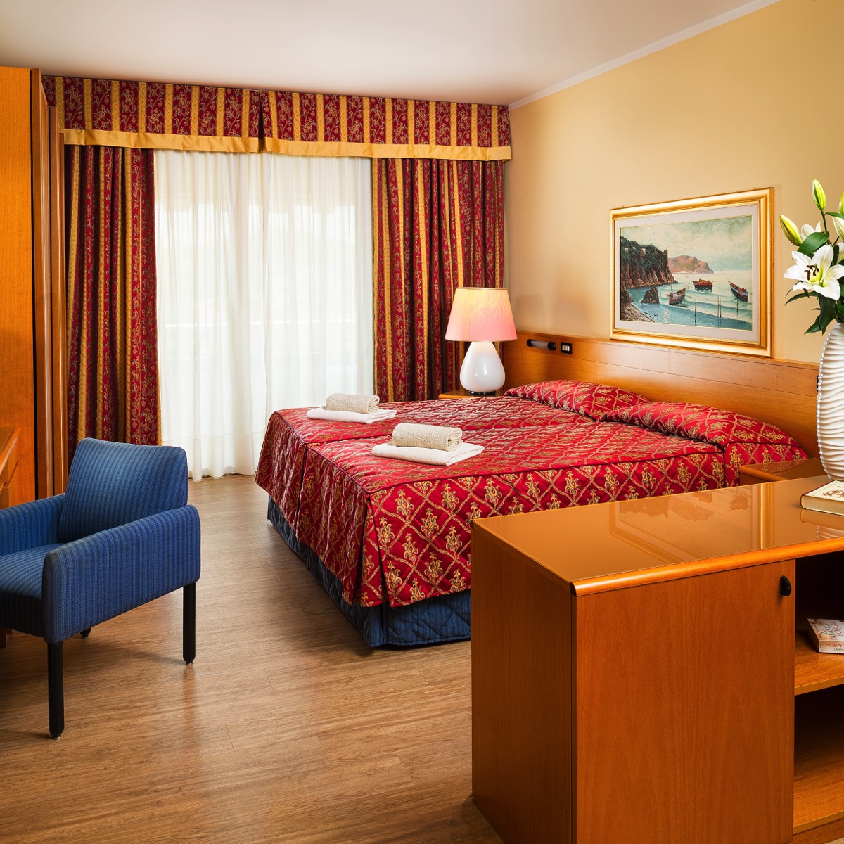 Camera d'hotel accogliente con letto matrimoniale, copriletto rosso, arredi in legno, lampada e tende su balcone luminoso.