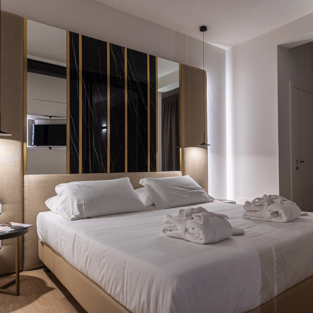 Camera hotel moderna con letto matrimoniale, testiera marmo nero-oro, accappatoi e luci a sospensione