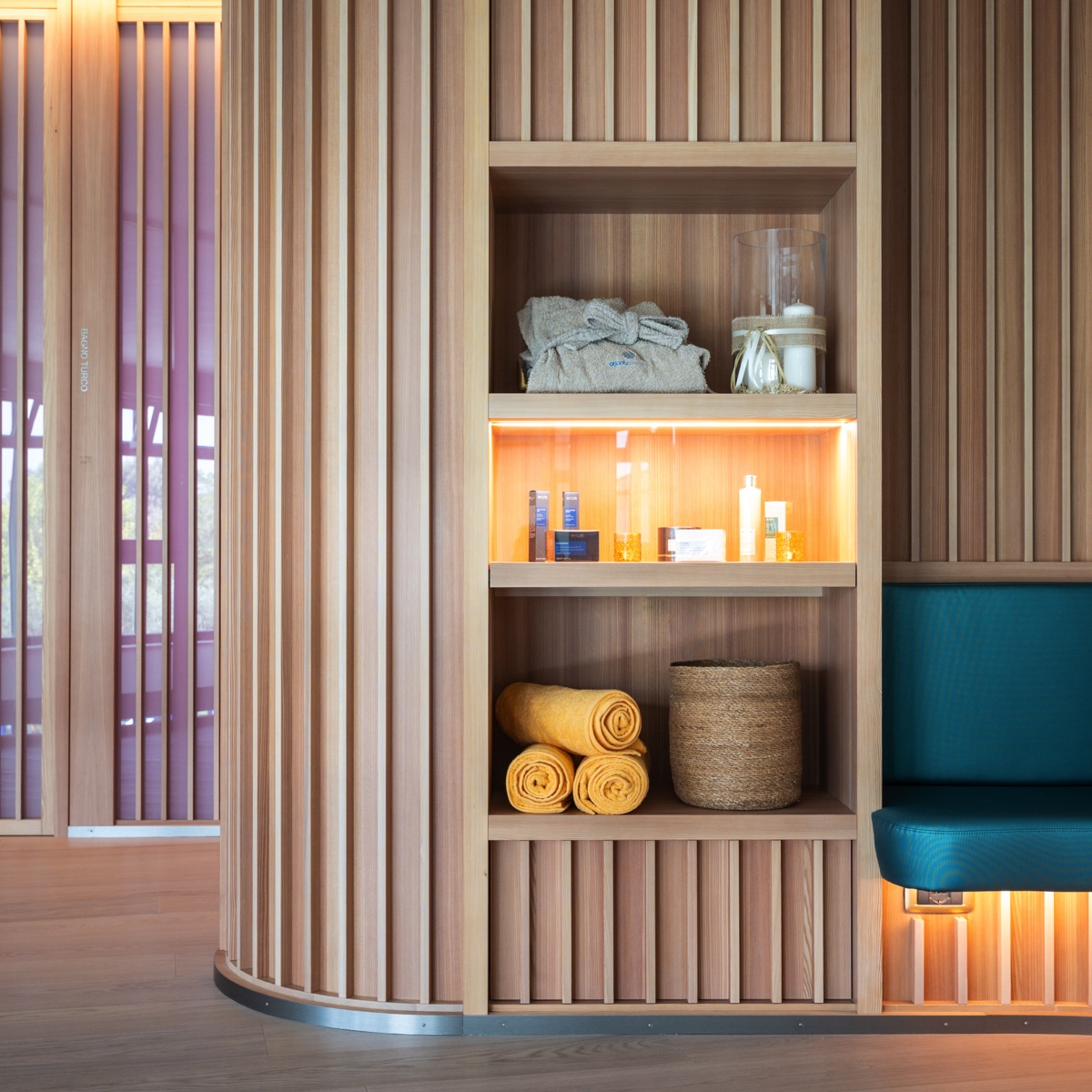 Scaffale spa in legno con accappatoio, prodotti skincare, asciugamani gialli e panca turchese, luce calda wellness.