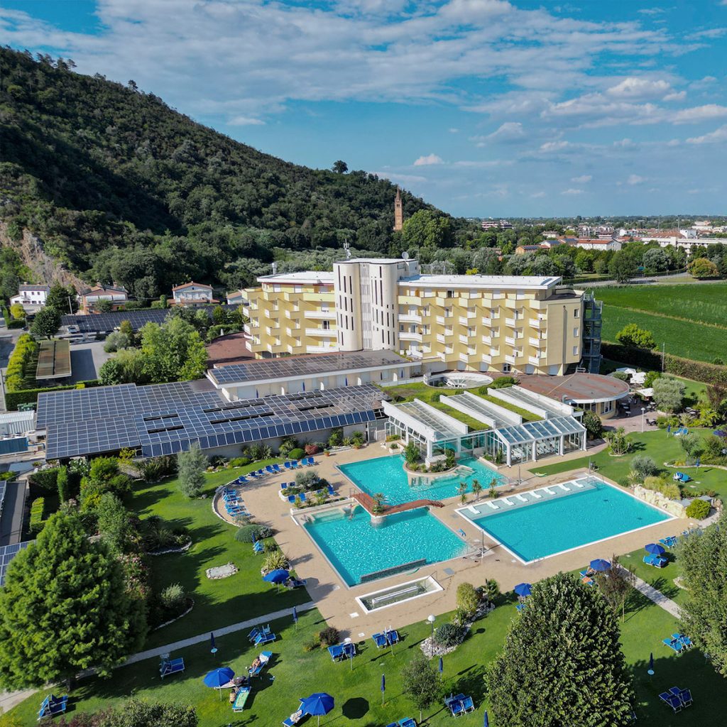 Vista aerea di resort termale con piscine termali esterne, giardini curati e pannelli solari ai piedi delle colline.