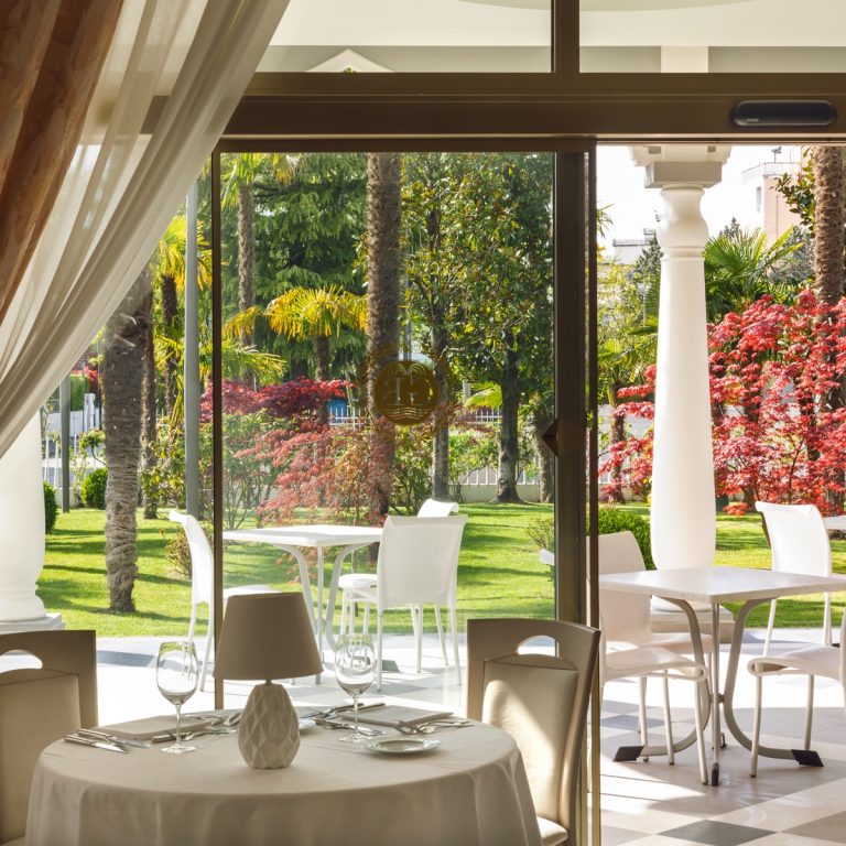 Ristorante elegante con tavolo apparecchiato e veranda luminosa, vista su giardino curato con palme e aceri rossi.