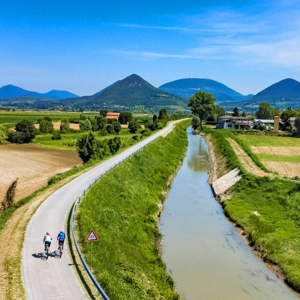 Ciclisti su pista ciclabile lungo canale in campagna veneta, con Colli Euganei e cielo azzurro sullo sfondo