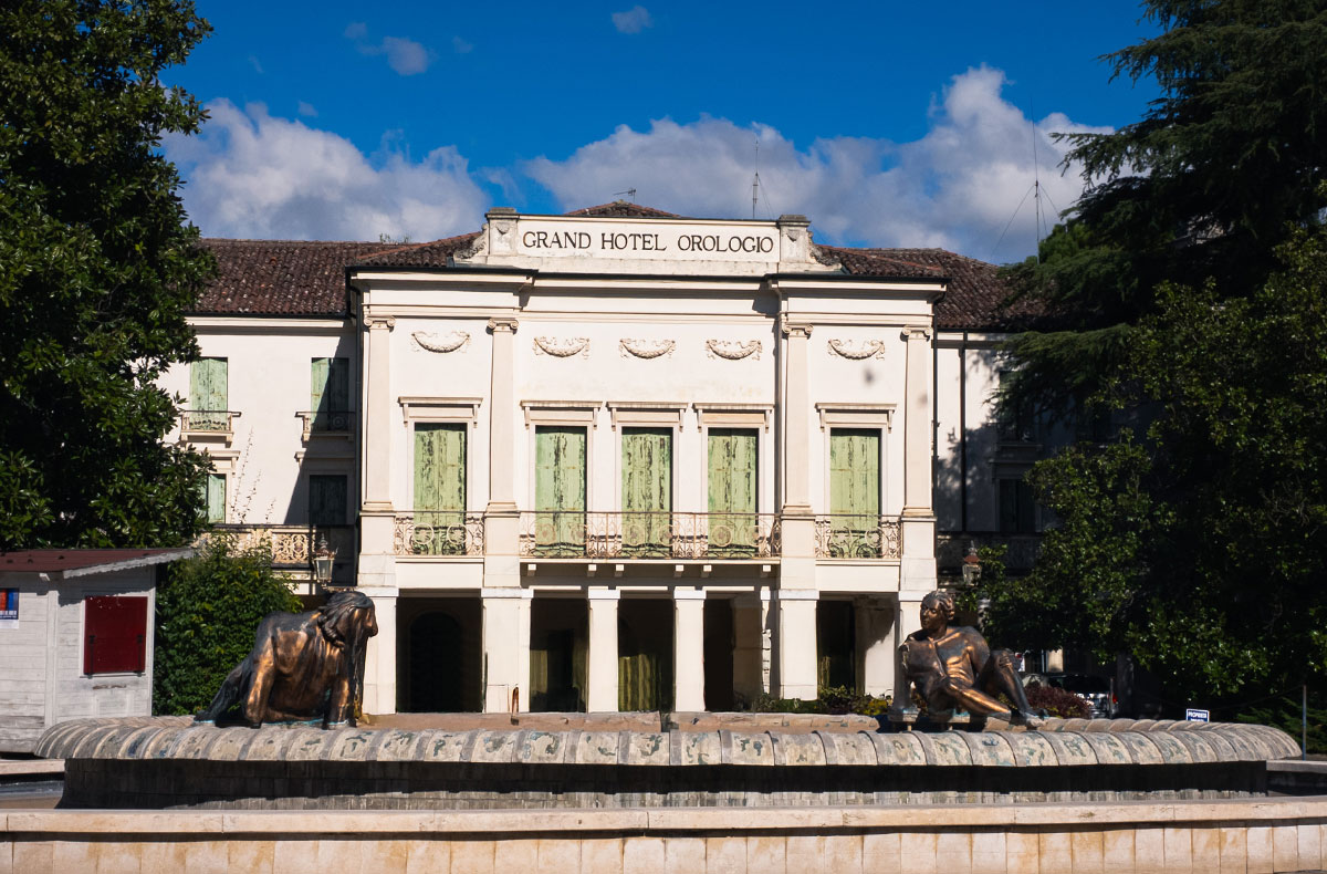 Facciata del Grand Hotel Orologio con fontana e statue bronzee in primo piano, cielo blu e alberi ai lati in Italia