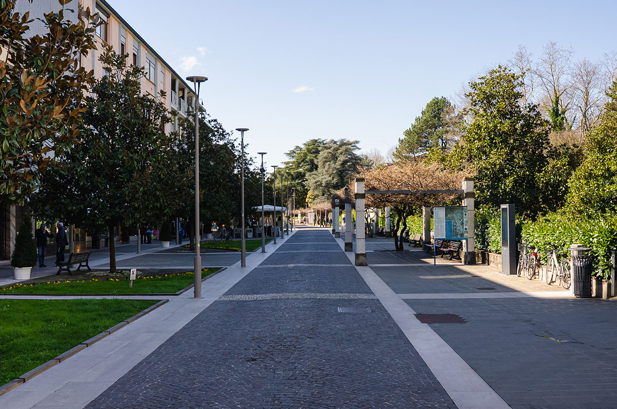 Viale pedonale alberato in centro città, pavé con panchine, lampioni e caffè, biciclette parcheggiate in giornata di sole.