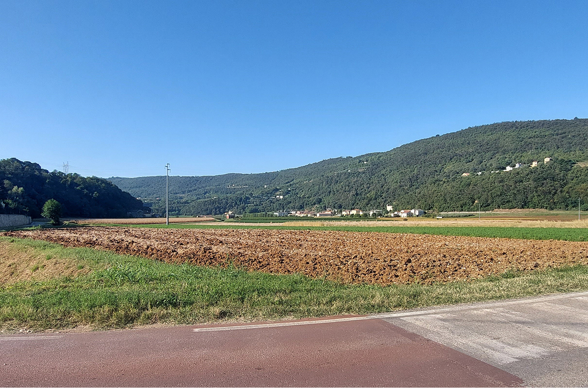 Campo arato in campagna con colline verdi, case sparse e cielo azzurro; paesaggio rurale estivo panoramico.