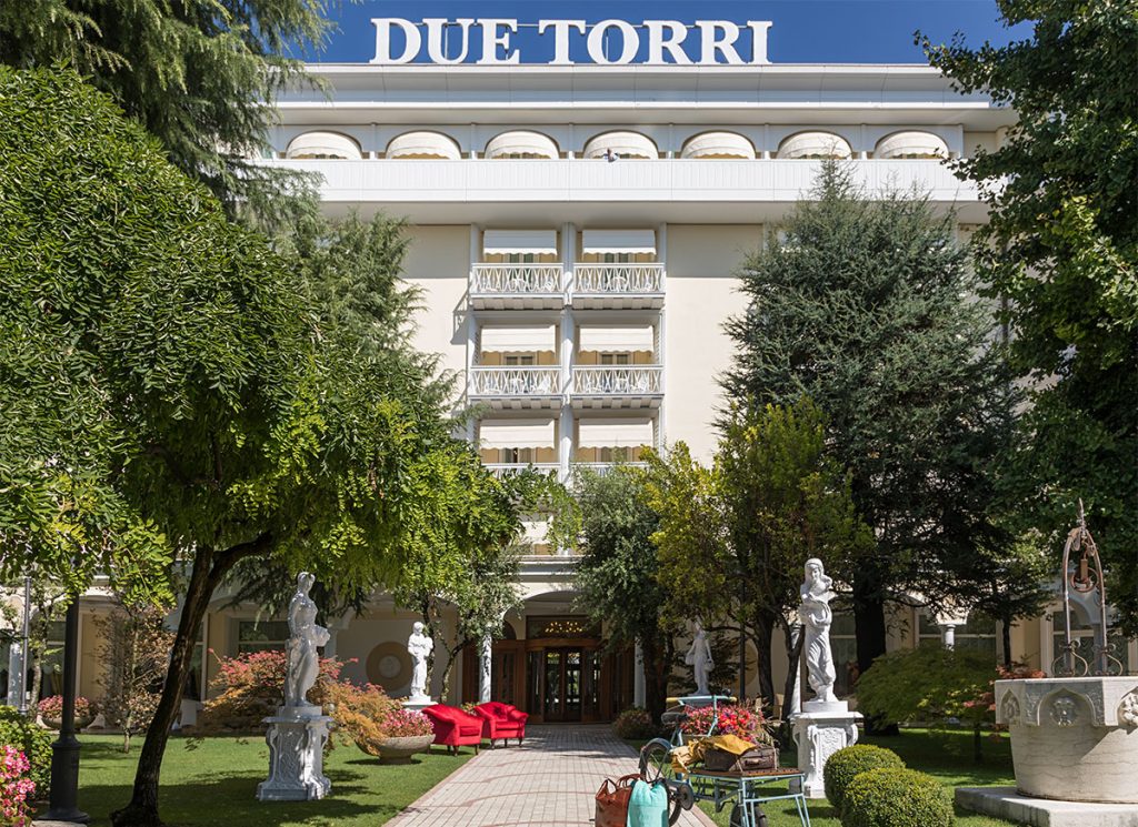 Hotel Due Torri: facciata elegante con giardino curato, statue classiche e ingresso con sedute rosse.
