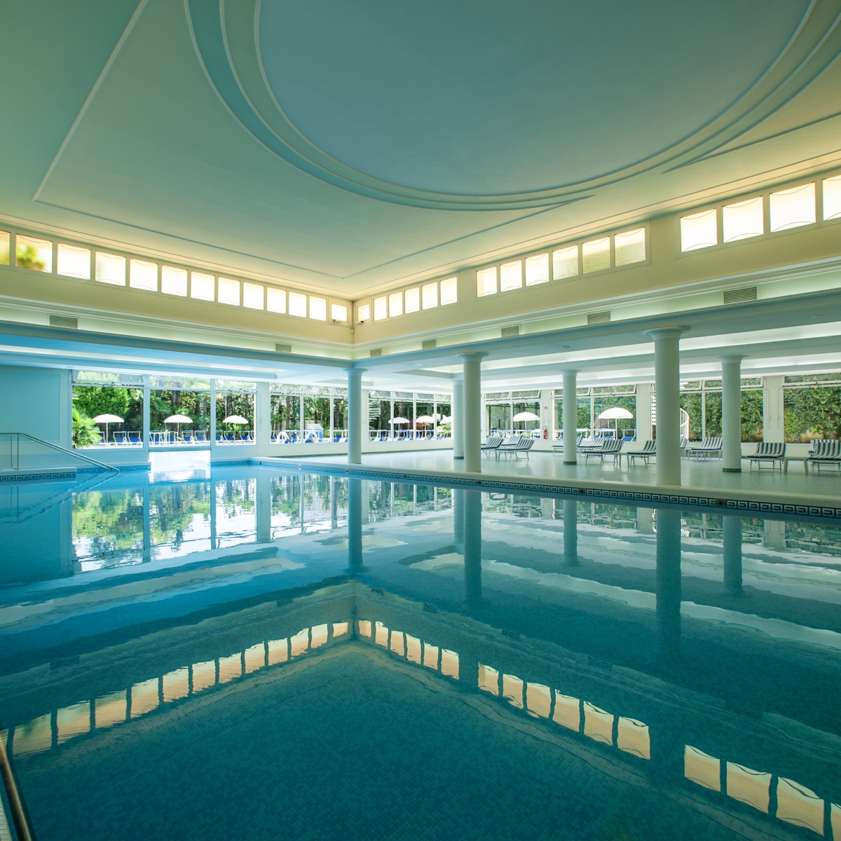 Piscina interna luminosa con colonne, ampie vetrate sul giardino e lettini relax in hotel spa di lusso.