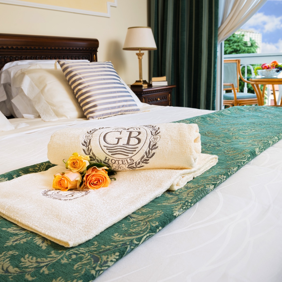 Elegante camera d'hotel: letto rifatto, asciugamani GB Hotels con rose, balcone con tavolino e luce naturale.