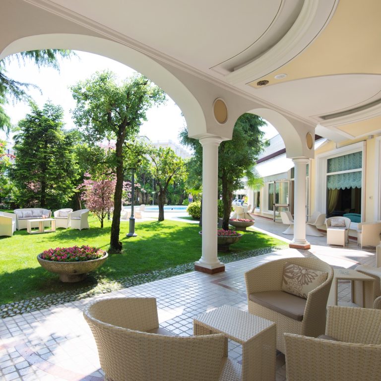 Elegante veranda d’hotel con archi e colonne, salottini in rattan, giardino curato con fiori e piscina sullo sfondo.