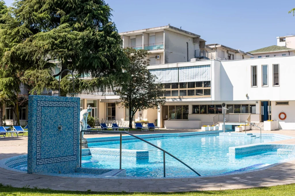 Hotel con piscina esterna a mosaico, sdraio e giardino alberato, giornata soleggiata con statua classica